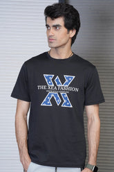 A male model wearing a black crew neck t-shirt with a graphic design in the center, standing in front of a metallic background.